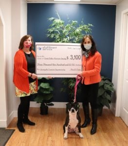 Two women stand on either side of a large check representing the donation LPC is making to the Great Lakes Humane Society. Patty Cake, the dog, is sitting in between them. Both humans are wearing face masks.